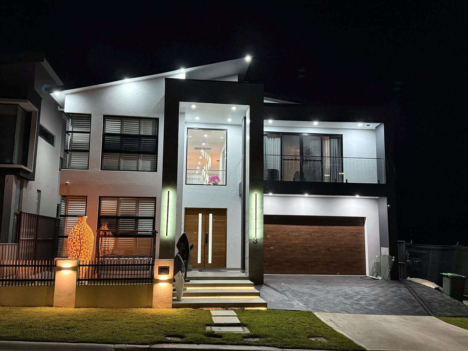 A modern double-storey home at night with a lit glazed entry, a timber double front door and a timber garage door