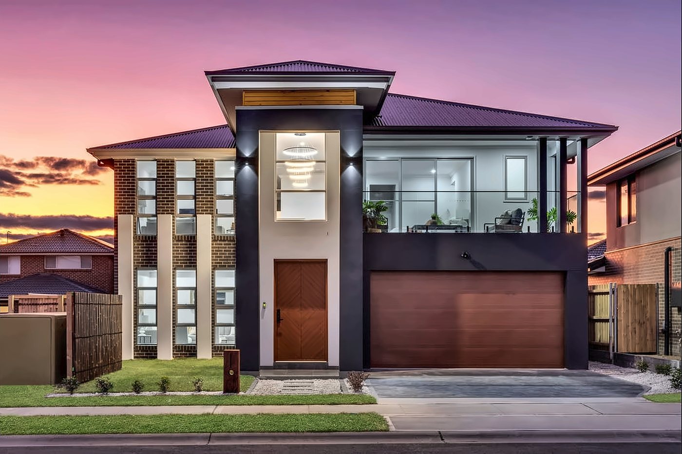 A double-storey home at sunset with a charcoal entry portal, a herringbone timber front door and a chandelier in the void
